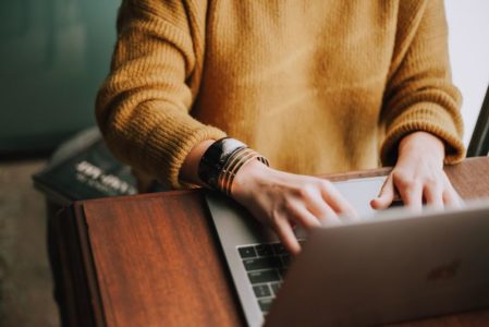 typing at laptop, yellow sweater, bracelets, wooden desk