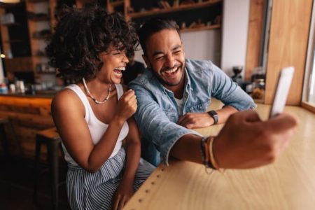 couple sitting together taking selfie, laughing, curly hair, bracelets, white tank top, blue shirt